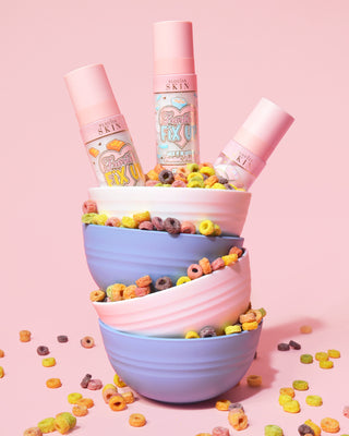 Stack of colourful bowls filled with cereal pouring out and three bottles of the P. Louise Frosted Fix Uo Blurring Setting Sprays in the top bowl on a pink background