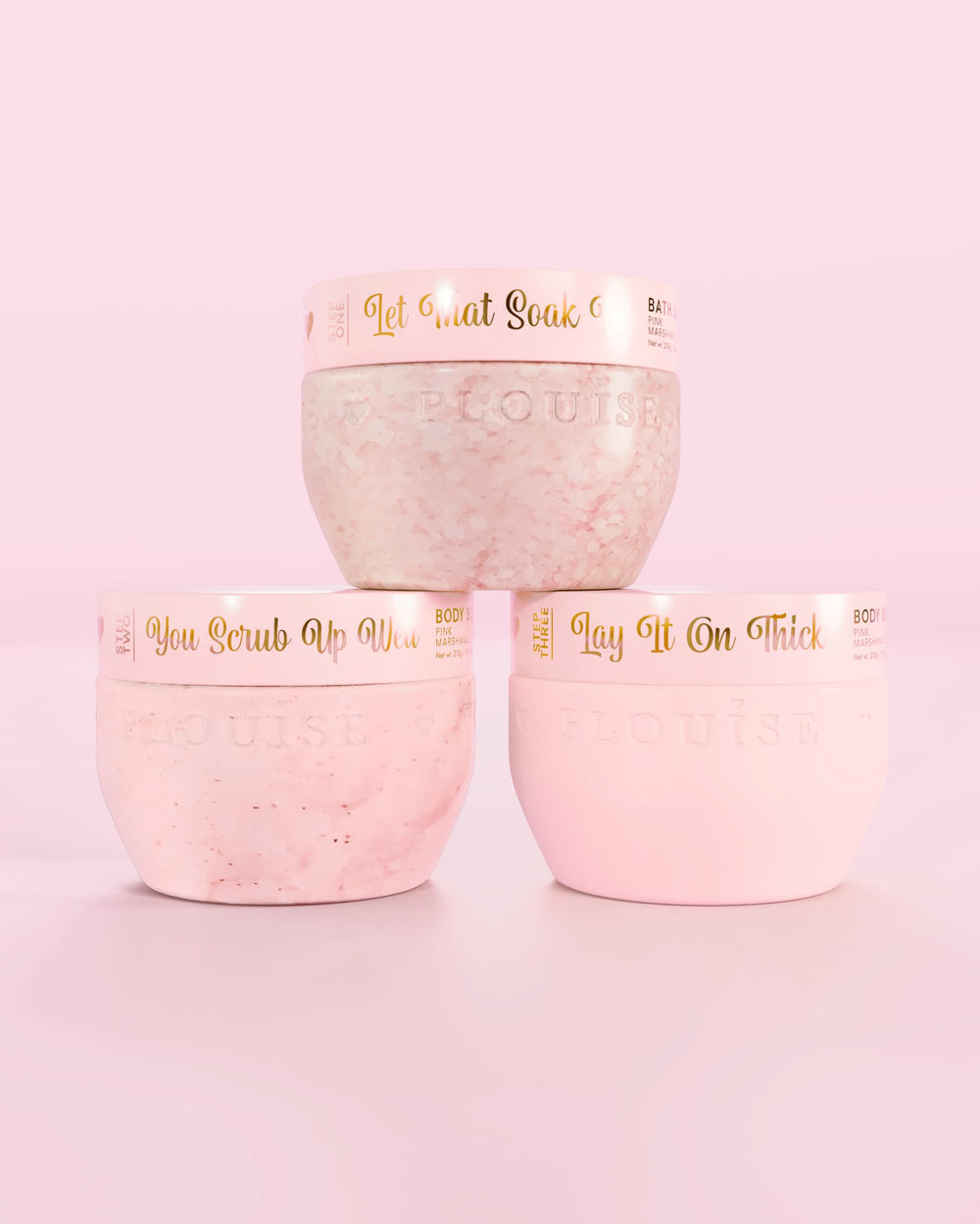 Trois contenants roses P.Louise Hop Into Self-Care Pink Marshmallow contenant des sels de bain, un gommage corporel et du beurre corporel avec un texte doré sur fond rose