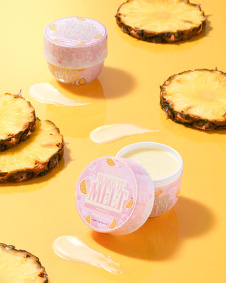 Two small jars of P. Louise Make It Melt Makeup Removing Cleansing Balm in Pineapple scent with a pink lid, surrounded by pineapple slices on a yellow background.