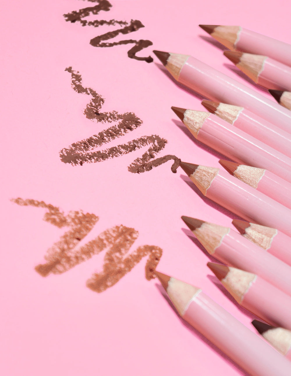 Une collection de crayons à lèvres aiguisables définition ultime P.Louise dans une variété de nuances nude, disposés sur un fond rose avec quelques lignes du produit dessinées sur la surface.
