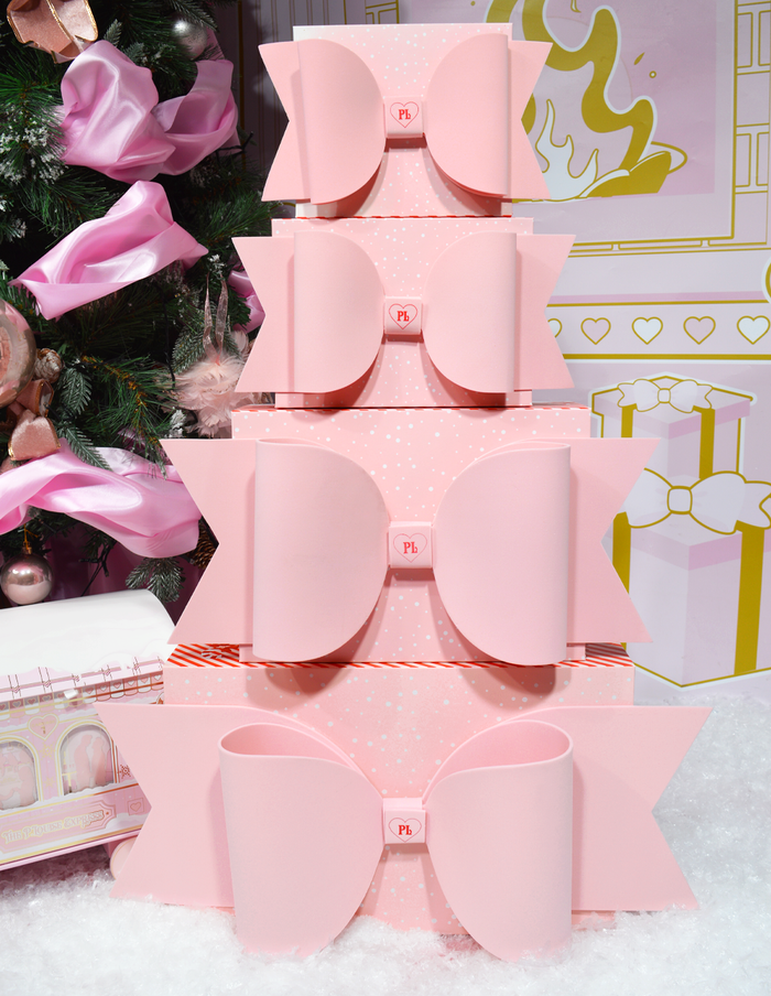 Pile de boîtes cadeaux roses P.Louise avec nœuds exposées sur fond de décorations sur le thème de Noël.