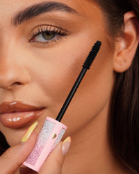 Close-up of a P.Louise Cry Baby mascara wand held near a person's eye with a pink glittery mascara tube.