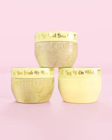 Trois contenants jaunes P.Louise Hop Into Self-Care Starwberry Shortcake contenant des sels de bain, un gommage corporel et du beurre corporel avec un texte doré sur fond rose