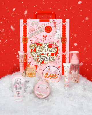 A festive decorative suitcase with Wrapped & Ready on it surrounded by P.Louise skincare products in hot toddy scent covered in snow