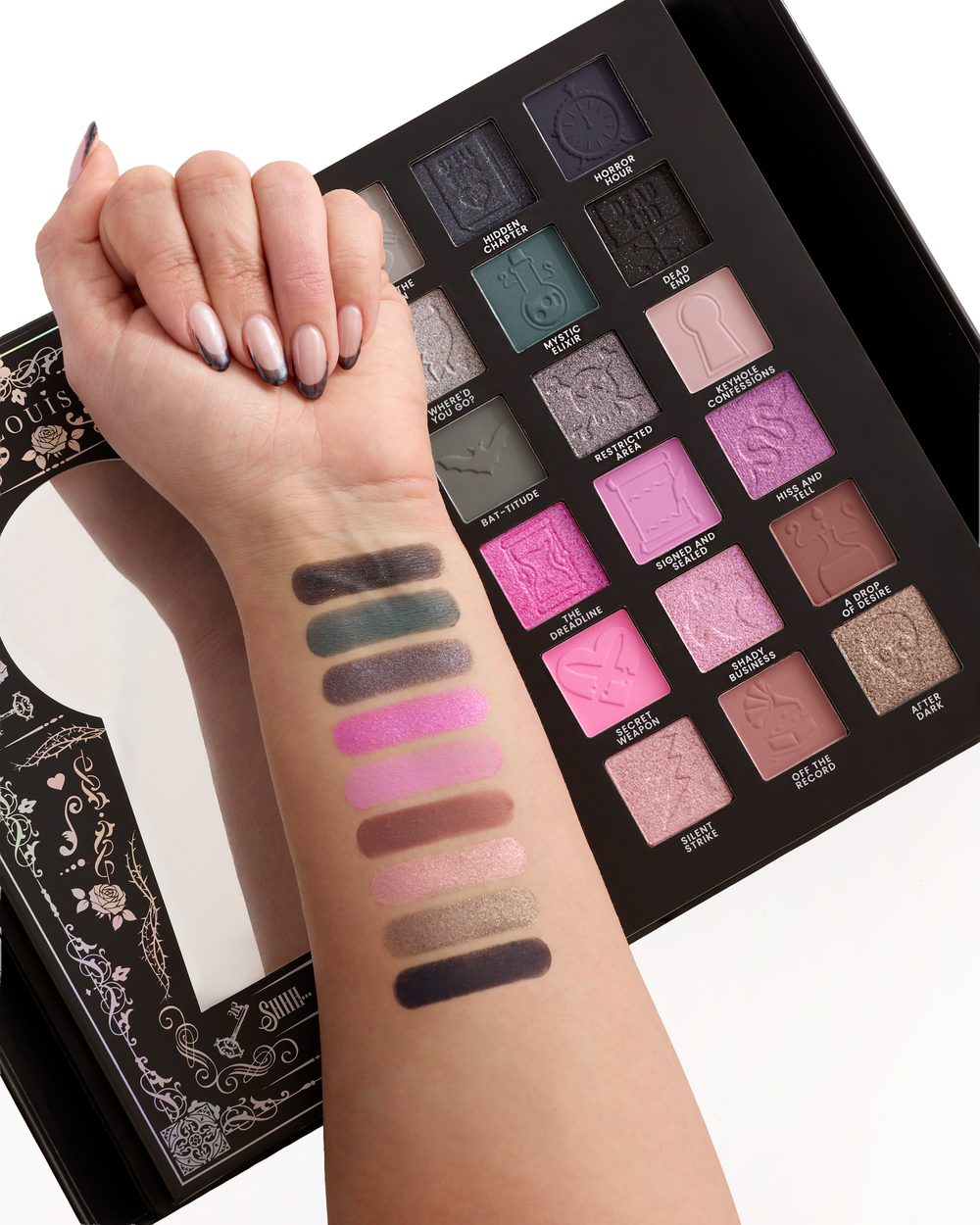A close-up of a halloween eyeshadow palette with an arm facing upwards next to it with vertical eyeshadow swatches painted on the forearm. The palette contains multiple shades including dark gray, teal, pink, purple, brown, and gold, with embossed script lettering on the pans and a decorative visible on the left.