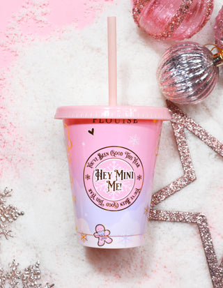 A pink P.Louise reusable plastic cup with a festive design and text reading 'Hey Mini Me' on the front. The cup includes a lid and a straw, and is presented on a white surface with decorative elements around it.