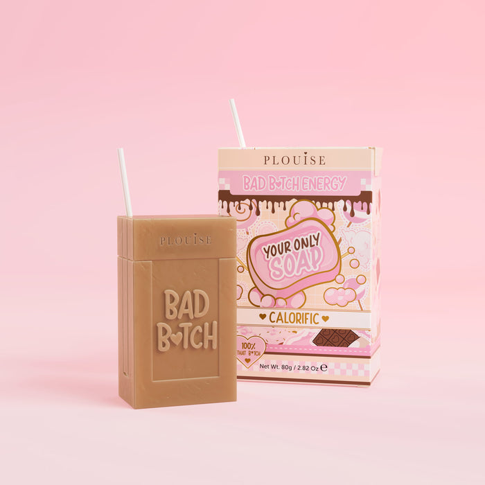 Un pain de savon marron sur lequel est gravé « Bad Bitch », accompagné d'un emballage de boîte de jus coloré portant l'inscription « Calorific », présenté sur fond rose.