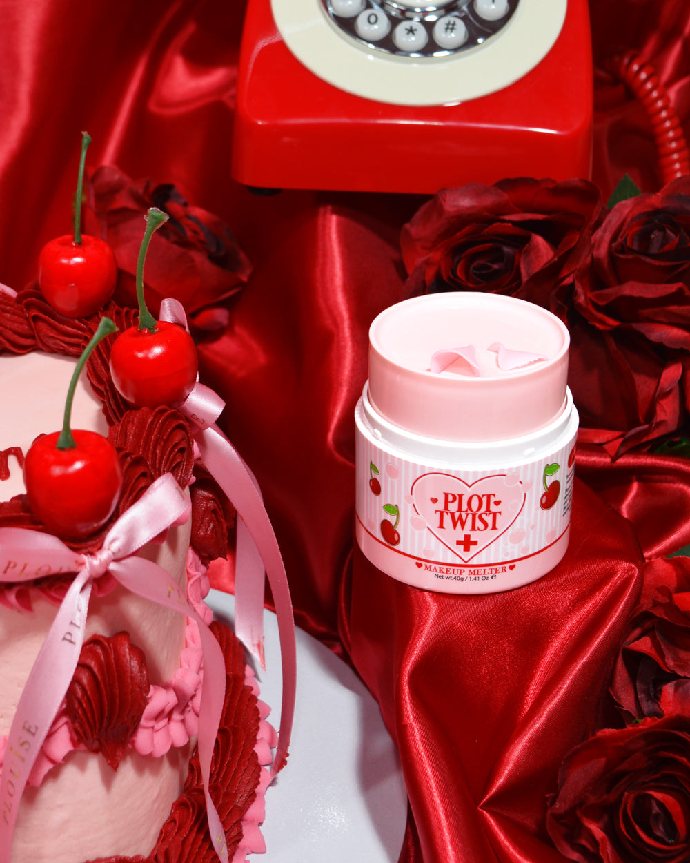 Un récipient de baume nettoyant de maquillage P.Louise Plot Twist Make It Melt affiché sur une surface en tissu rouge avec des éléments décoratifs dont une cerise et un téléphone rouge vintage en arrière-plan.